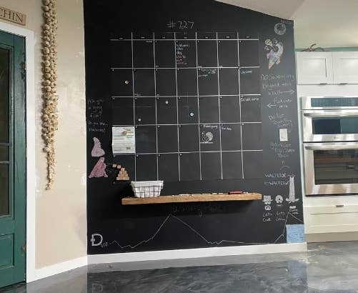 A large chalkboard wall in a kitchen with a calendar grid, various drawings, motivational quotes, and a shelf holding a white basket. A green door is on the left