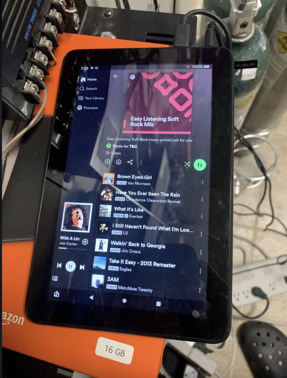 A tablet showing a Spotify playlist titled "Easy Listening Soft Rock Mix" is displayed on a cluttered surface, including wires and an orange Amazon box