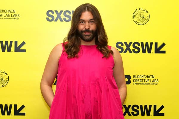 Jonathan Van Ness poses in a sleeveless, pink dress at the SXSW event, standing against a yellow backdrop with logos