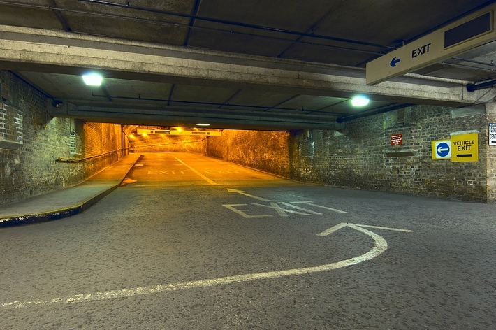 An empty underground parking garage with signs indicating the exit route. The walls are made of bricks and the floor has direction arrows. No people are present