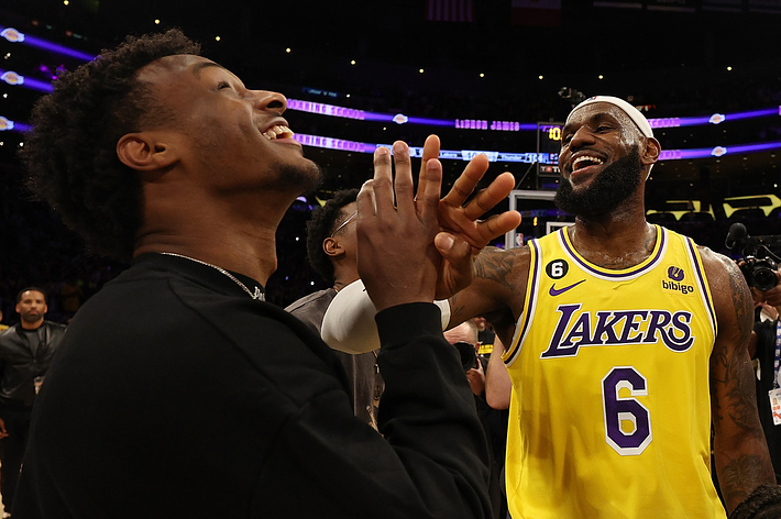 LeBron James in a Lakers jersey and his son Bronny James share a joyful moment on a basketball court, laughing and clasping hands