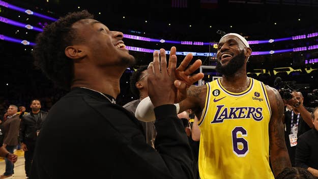 LeBron James, wearing a Lakers jersey, shares a laugh with Bronny James on a basketball court, surrounded by people and bright lights in the background