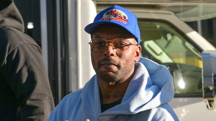 Man wearing a blue cap with &quot;Monaco&quot; text and a graphic, glasses, and a light-colored hoodie, posing outdoors near a vehicle