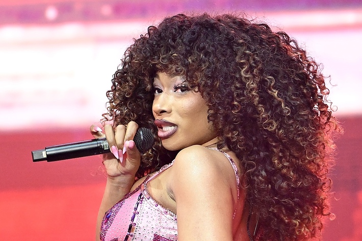 Megan Thee Stallion performs on stage, holding a microphone, with voluminous curly hair and wearing a sparkling outfit