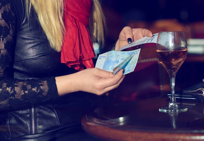 Person counting Euro banknotes at a table, with a glass of wine nearby. Their face is not visible, and they are wearing a leather jacket and red scarf