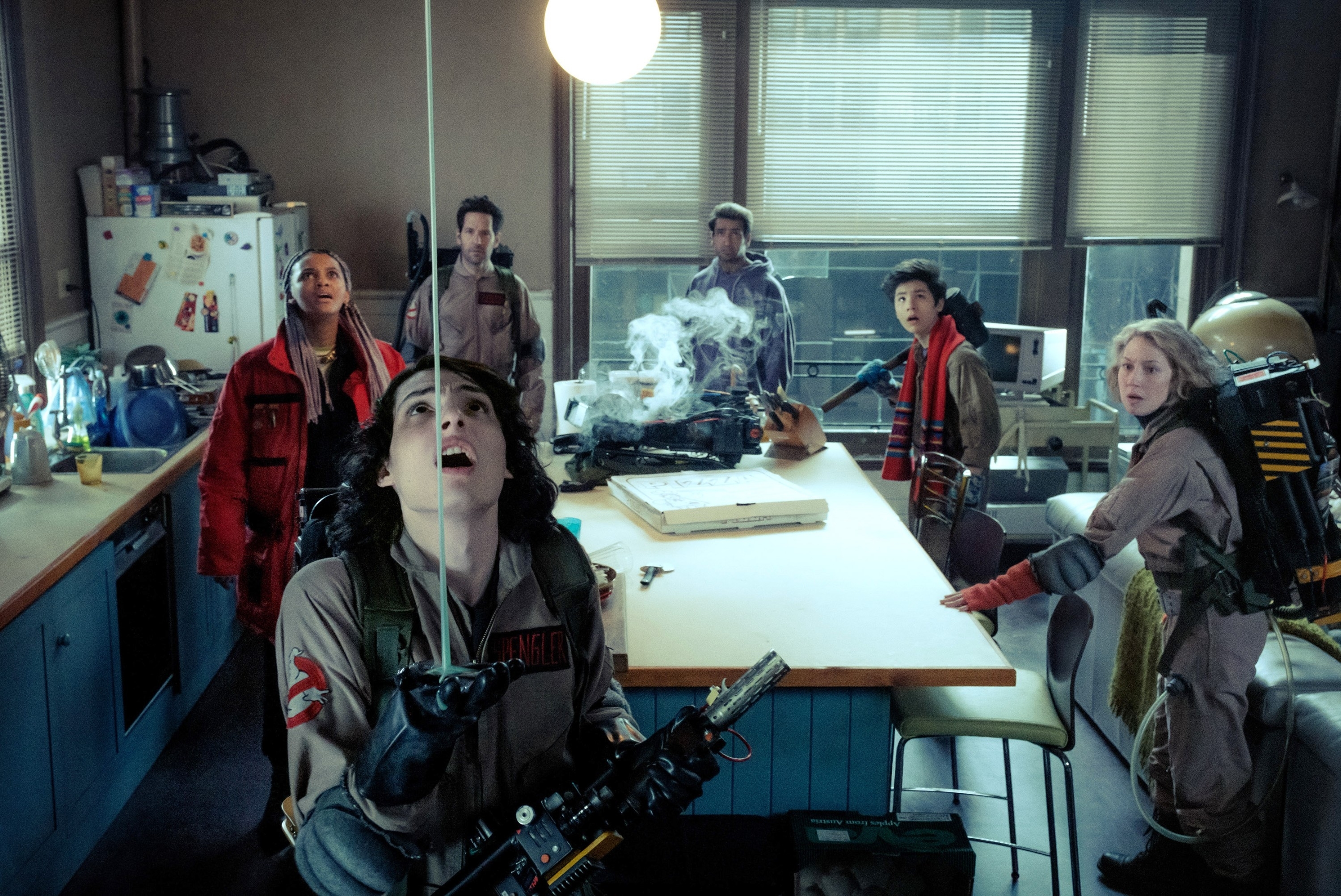 Finn Wolfhard, Mckenna Grace, and cast from &quot;Ghostbusters: Afterlife&quot; in a laboratory scene, dressed in ghostbuster uniforms, looking upward with gear in hand