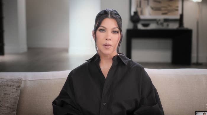 Kourtney Kardashian sits on a beige sofa in a minimalist room, wearing a black button-up shirt, with her hair styled in a sleek bun