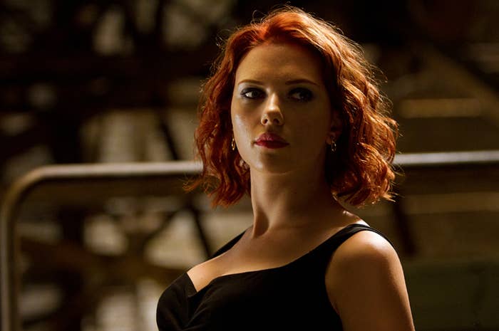 Scarlett Johansson, with short red hair, wearing a sleeveless black dress, is standing and looking to the side. Background is blurred