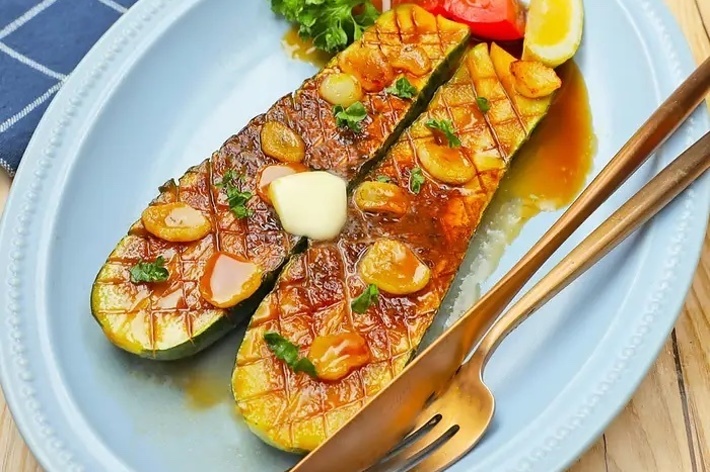野菜料理の写真。焼いたズッキーニが盛り付けられ、ニンニクのスライス、バター、パセリが添えられています。バックにはナイフとフォーク。