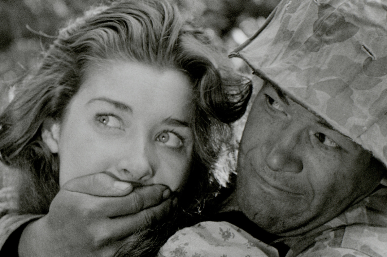 A man in military attire holds his hand over a woman's mouth. The woman looks worried. Names of people are unknown