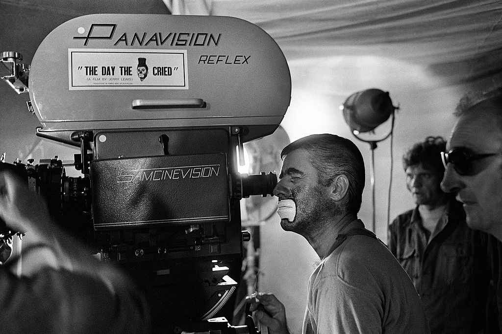 Several people are on a film set with a Panavision camera. One person with clown makeup is looking through the viewfinder