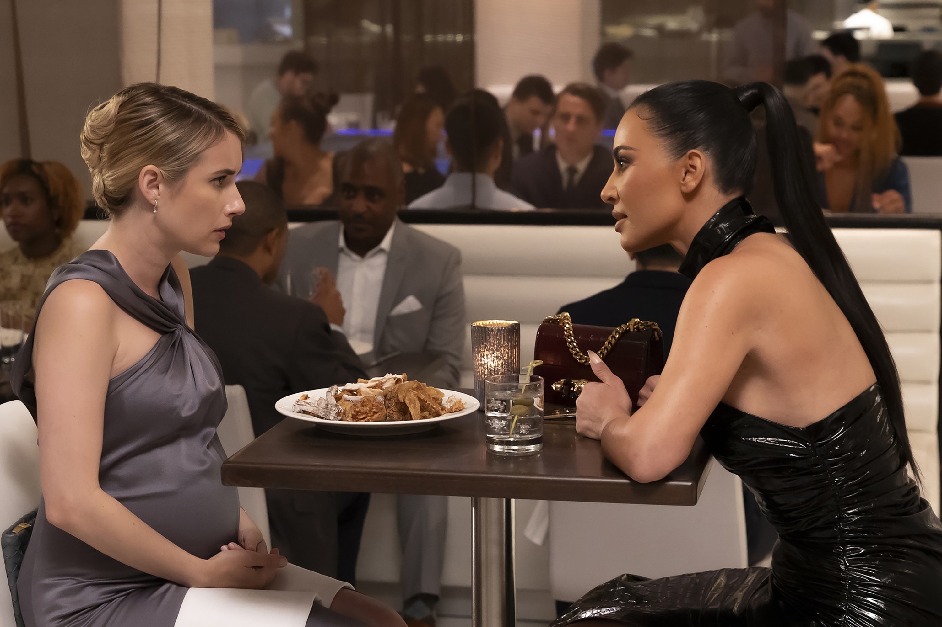 Emma Roberts and Kim Kardashian sit at a restaurant table, engaged in a serious conversation. Emma wears a dress, while Kim wears a black leather outfit