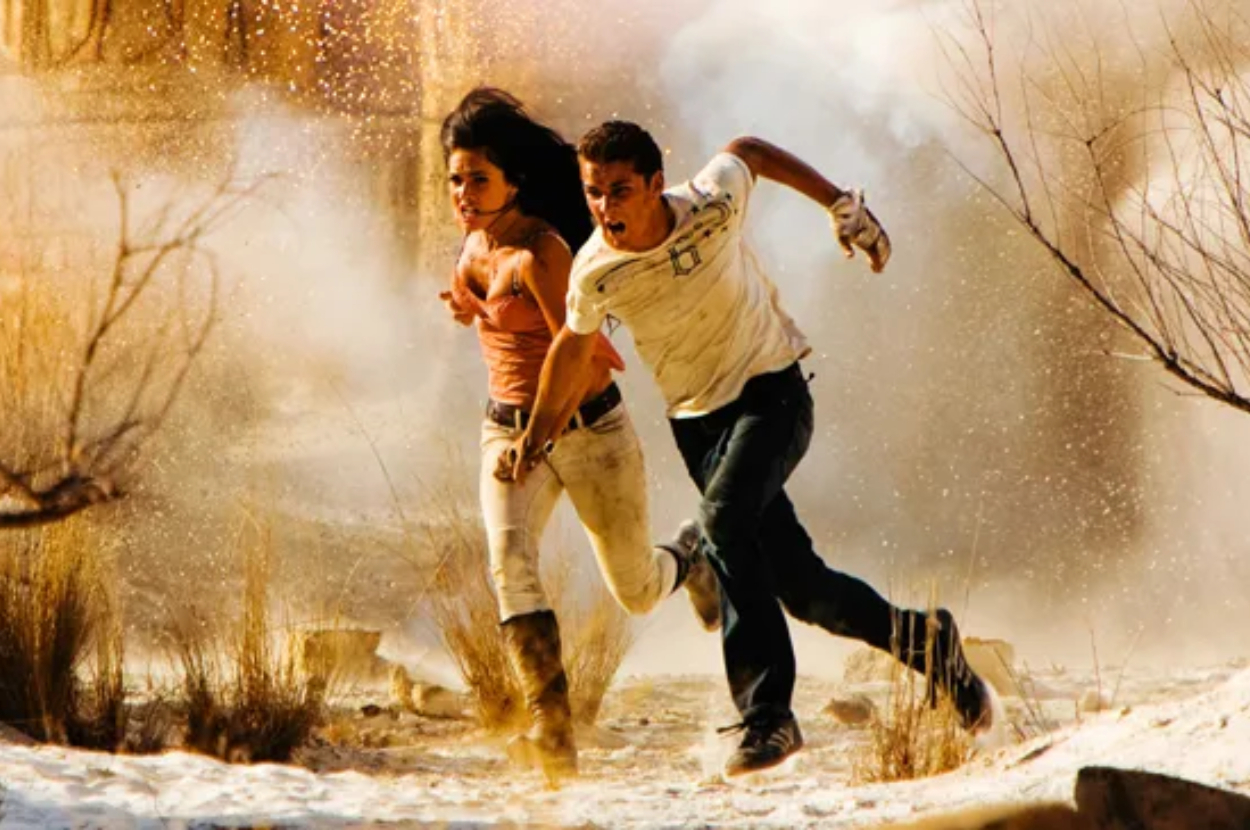 Megan Fox and Shia LaBeouf are running through an arid, dusty landscape with determined expressions, appearing to escape from an unseen threat