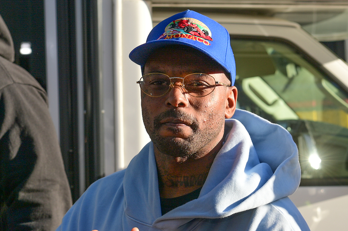 A man wearing a blue cap and glasses with a hooded sweatshirt stands outside