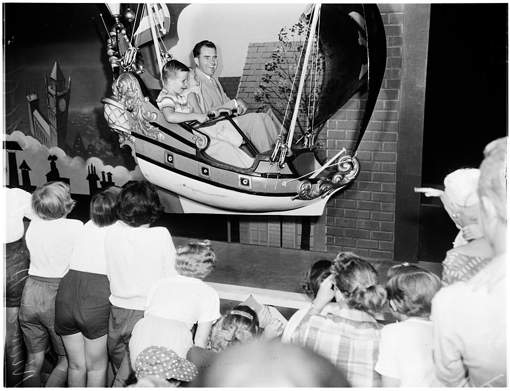 Walt Disney and a young girl riding a Peter Pan's Flight attraction as a crowd of children watches in awe
