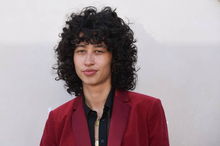 Person with curly hair wearing a stylish velvet blazer and a collared shirt, smiling slightly. The background is plain