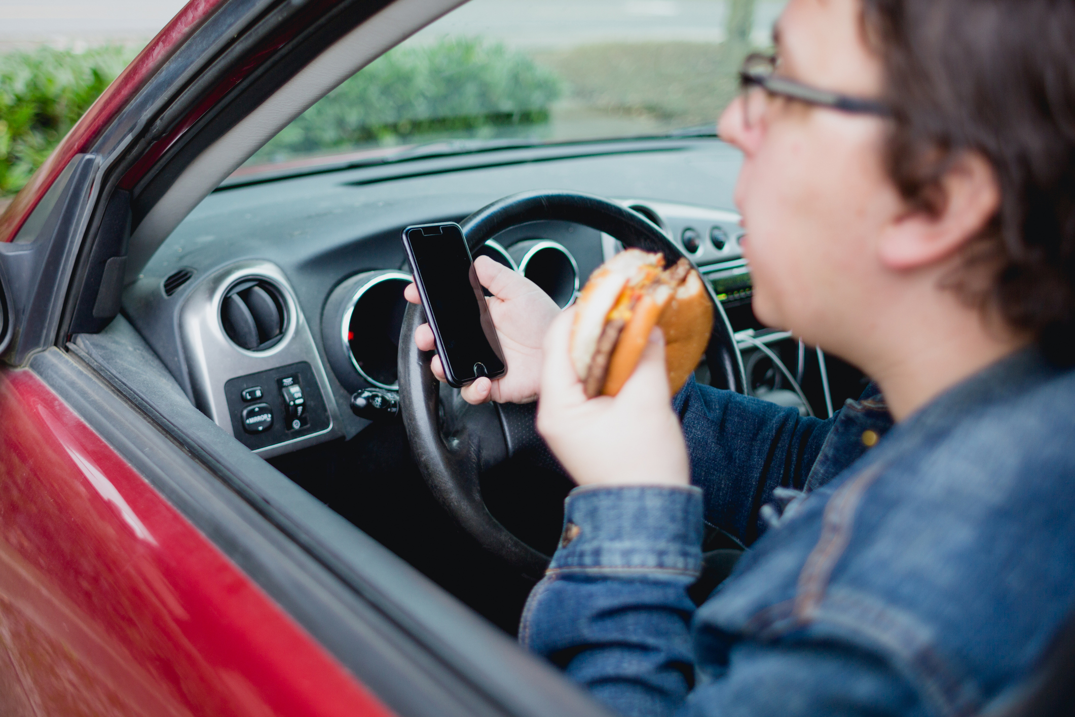 Person driving a car while holding a smartphone in one hand and a sandwich in the other