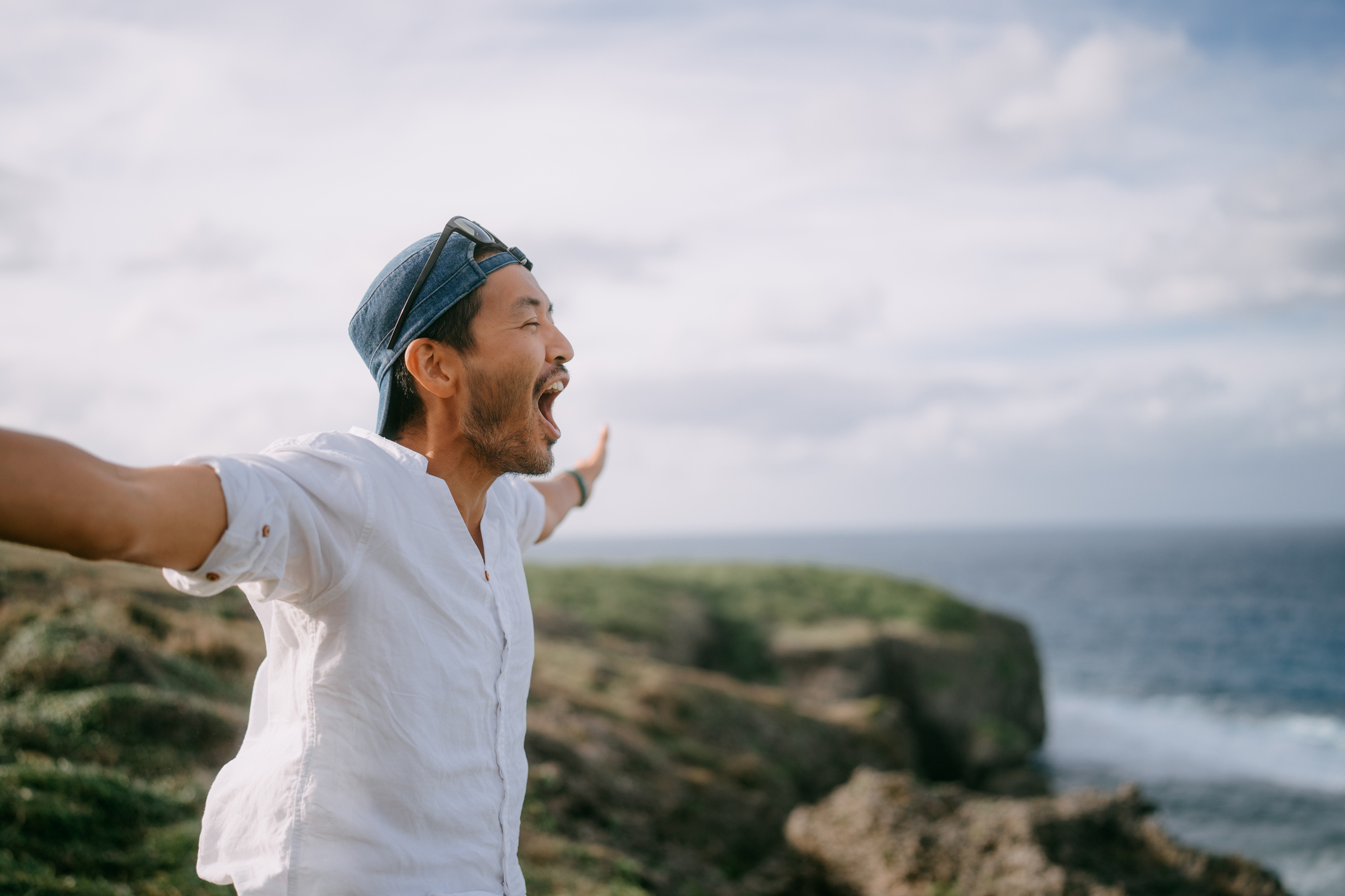 Man standing on a cliff near the ocean, arms outstretched, wearing a white shirt and a backwards cap