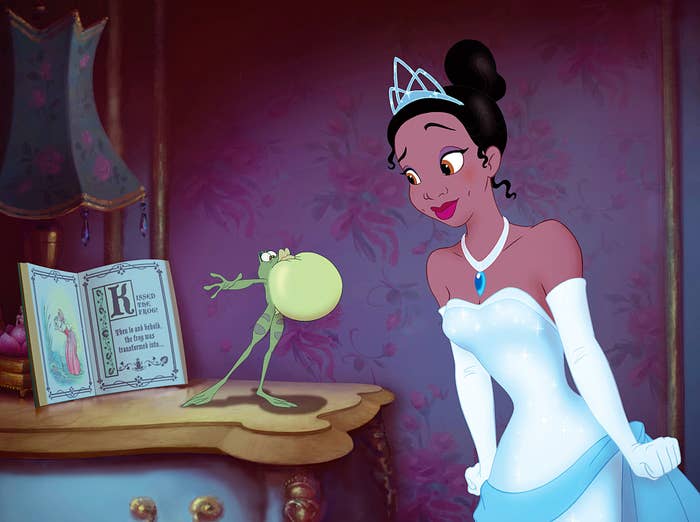 Tiana from The Princess and the Frog looks at frog Prince Naveen standing on a table next to a fairy tale book in a bedroom setting