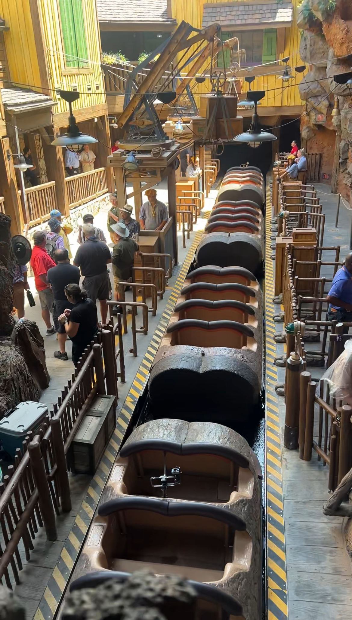 People are boarding a roller coaster ride at an amusement park. The setting has a rugged, Western theme with wooden structures and a mountain backdrop