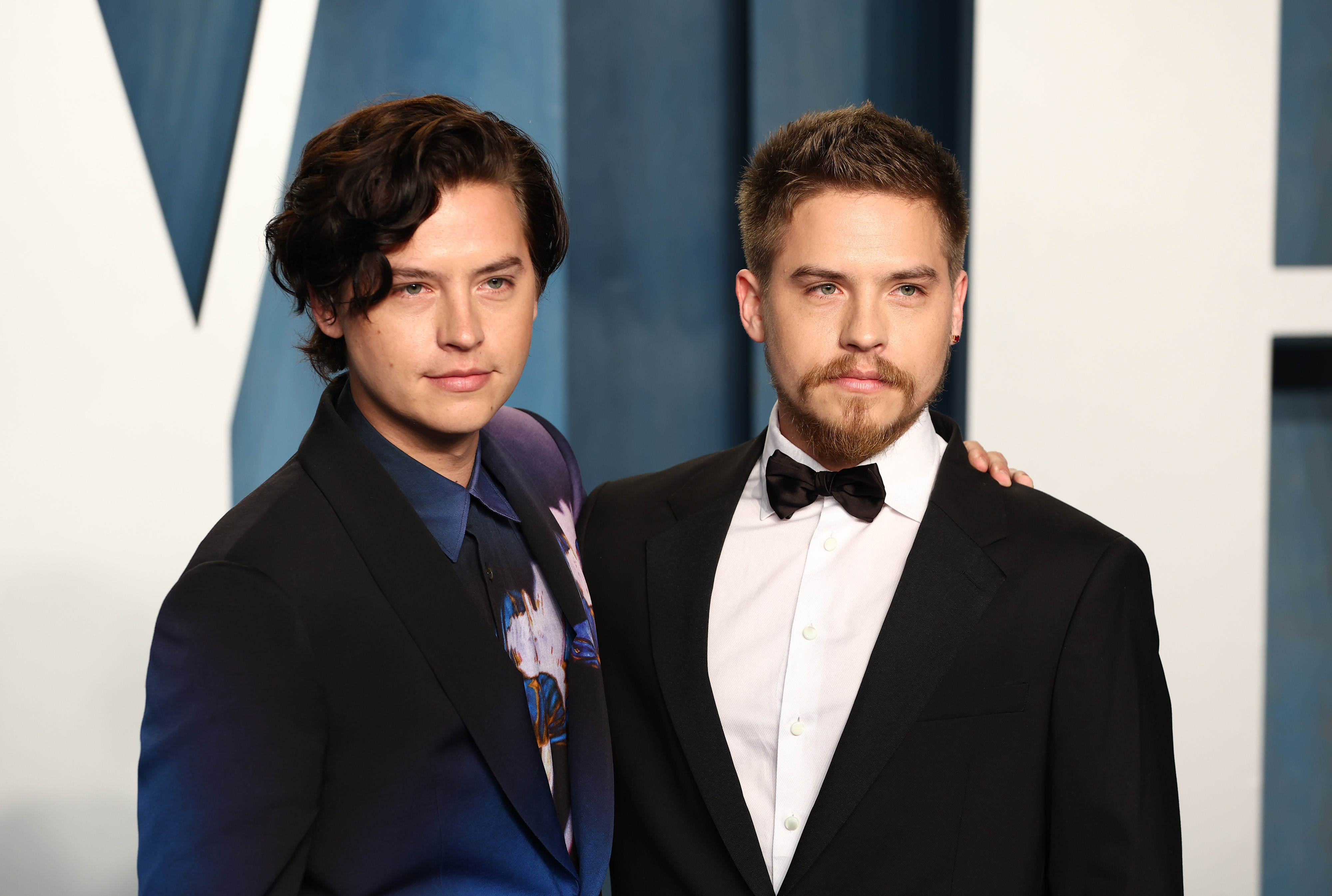 Cole Sprouse in a printed suit jacket stands next to Dylan Sprouse in a classic black tuxedo with a white shirt and black bow tie