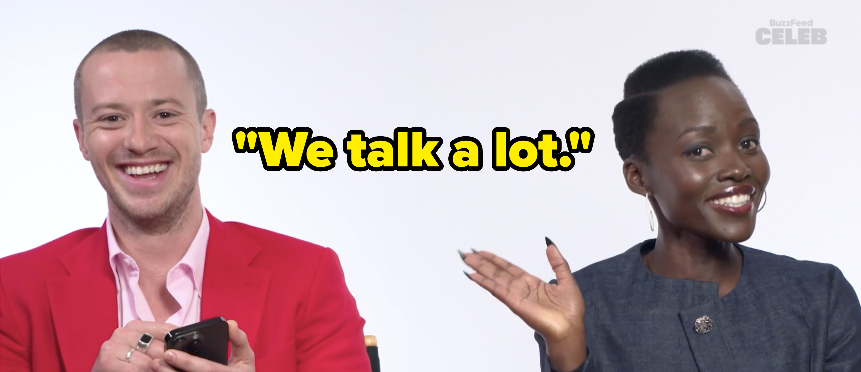 Lupita Nyong'o and an unidentified person are seated and smiling during a BuzzFeed Celeb segment. Lupita wears a dark blazer, and the other person is in a pink shirt and red jacket