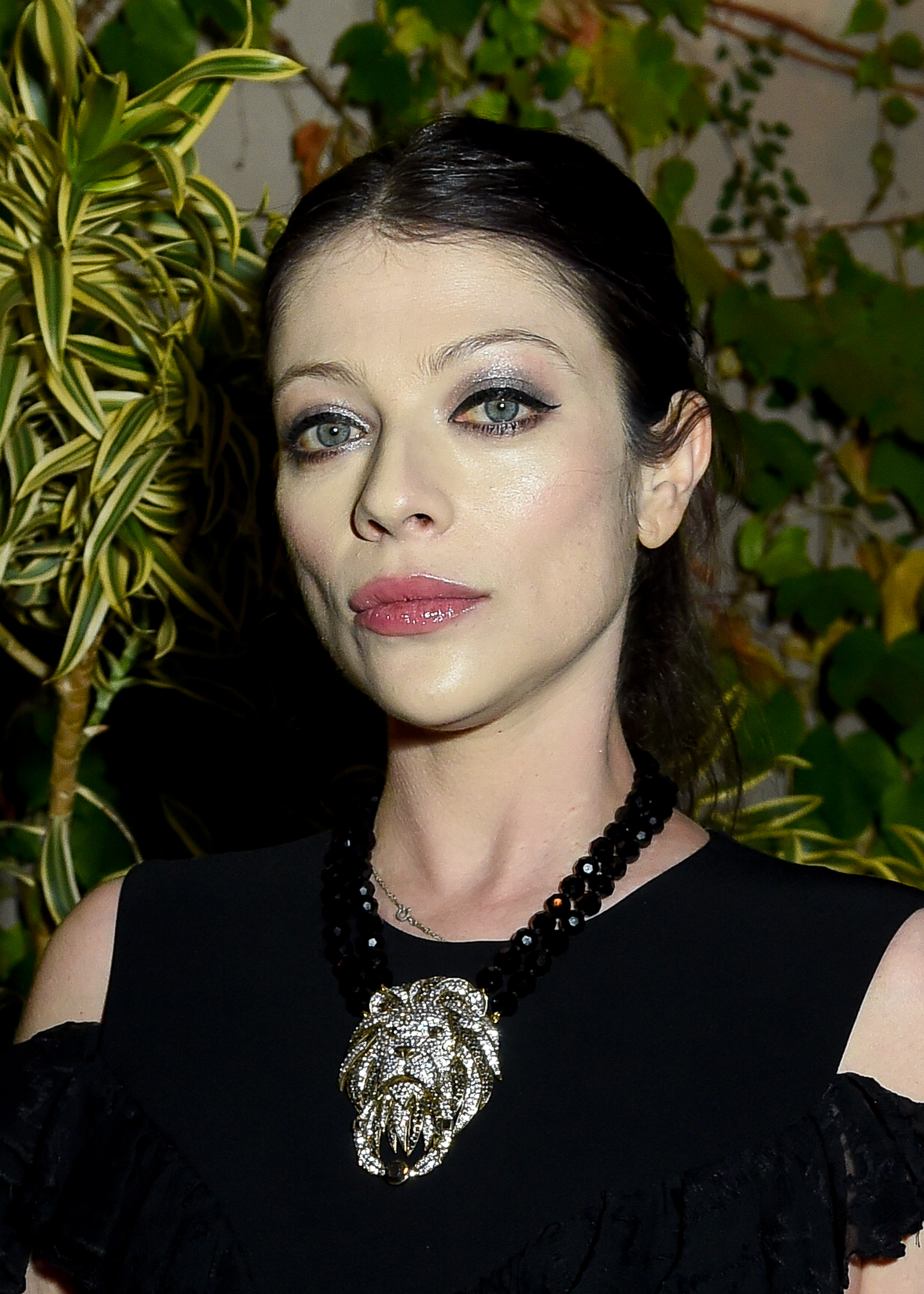 Michelle Trachtenberg is shown in a portrait wearing a black dress and a statement necklace with a large, ornate pendant