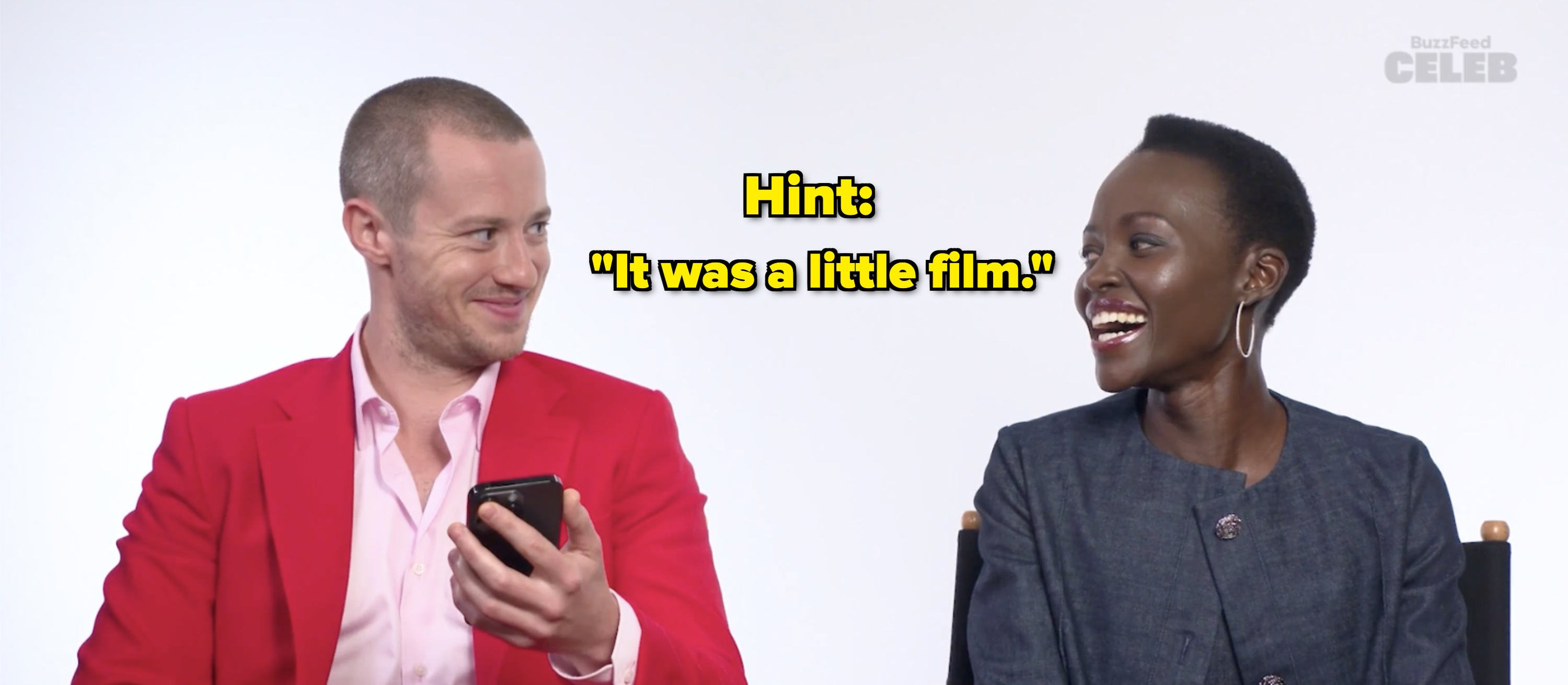 Joseph Quinn in a red blazer and Lupita Nyong'o in a dark blazer laughing during a BuzzFeed Celeb interview on a white background