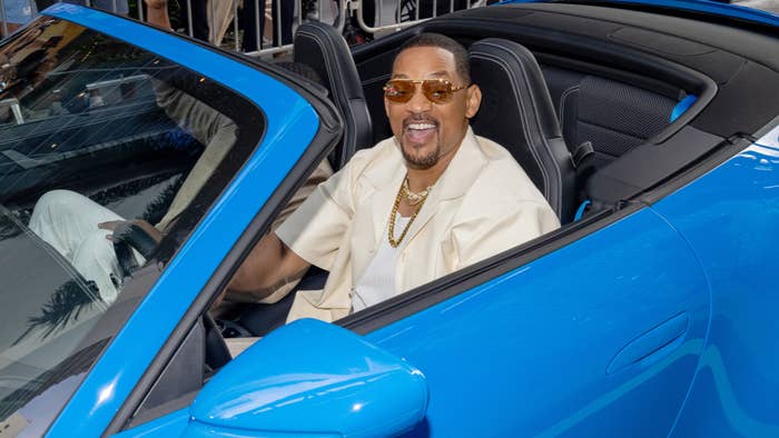 Will Smith sitting in a blue convertible car, smiling and wearing sunglasses, a beige suit jacket, and a gold chain