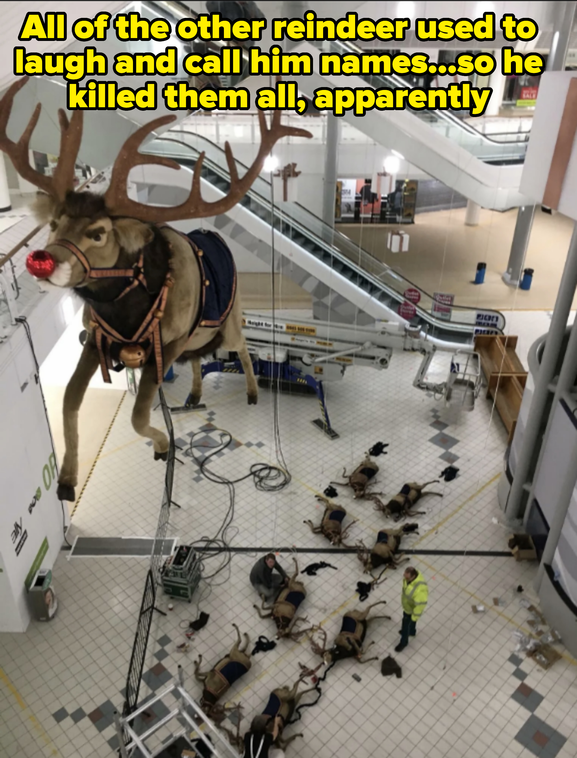 Large reindeer decorations hanging by ropes in a shopping mall; a crew is setting them up. Several people in yellow high-visibility jackets are visible