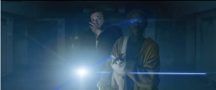 A man and a woman hold a cat while standing in a dimly-lit area, with a bright light shining towards them