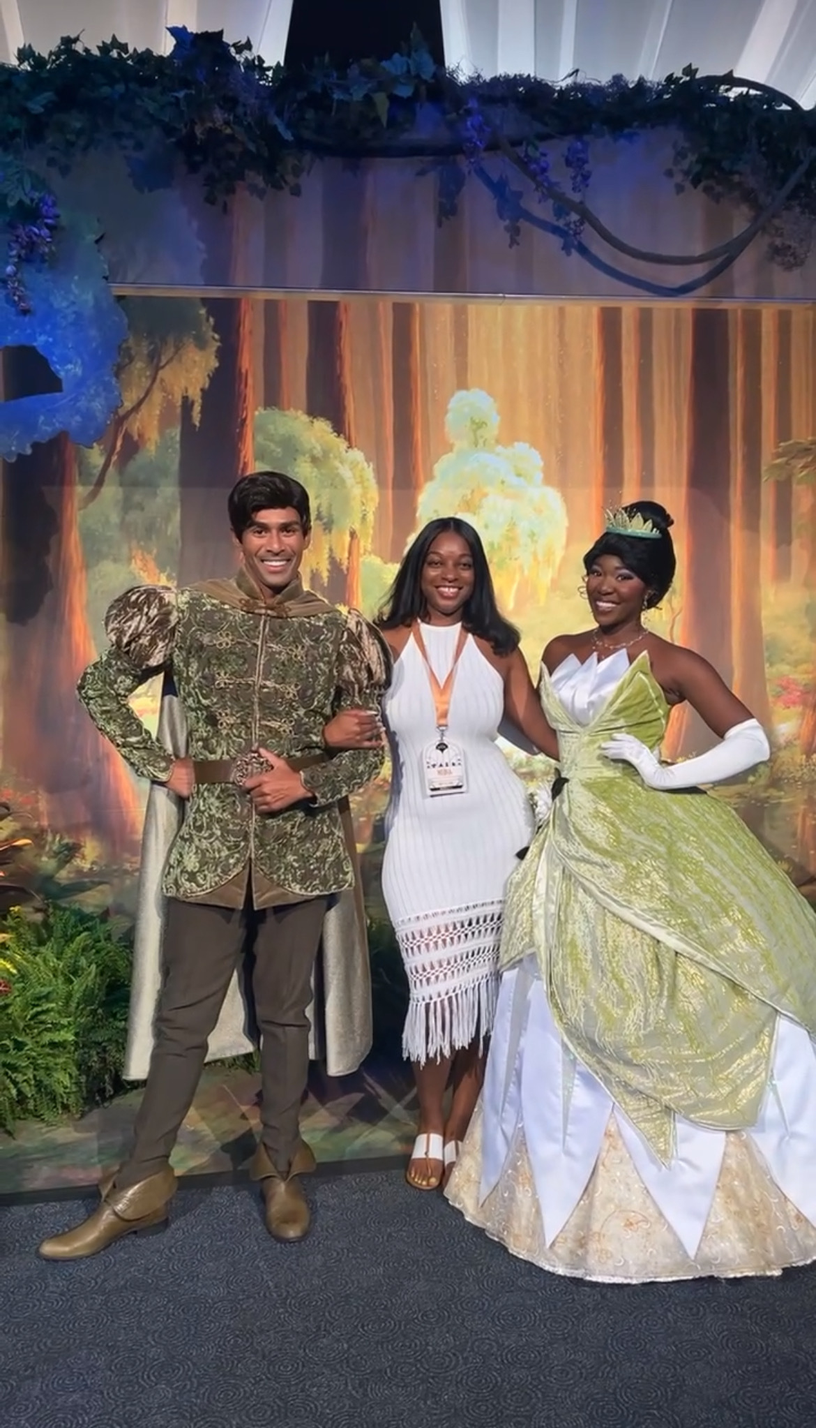 Two people dressed as Prince Naveen and Princess Tiana pose with a smiling guest at a themed event