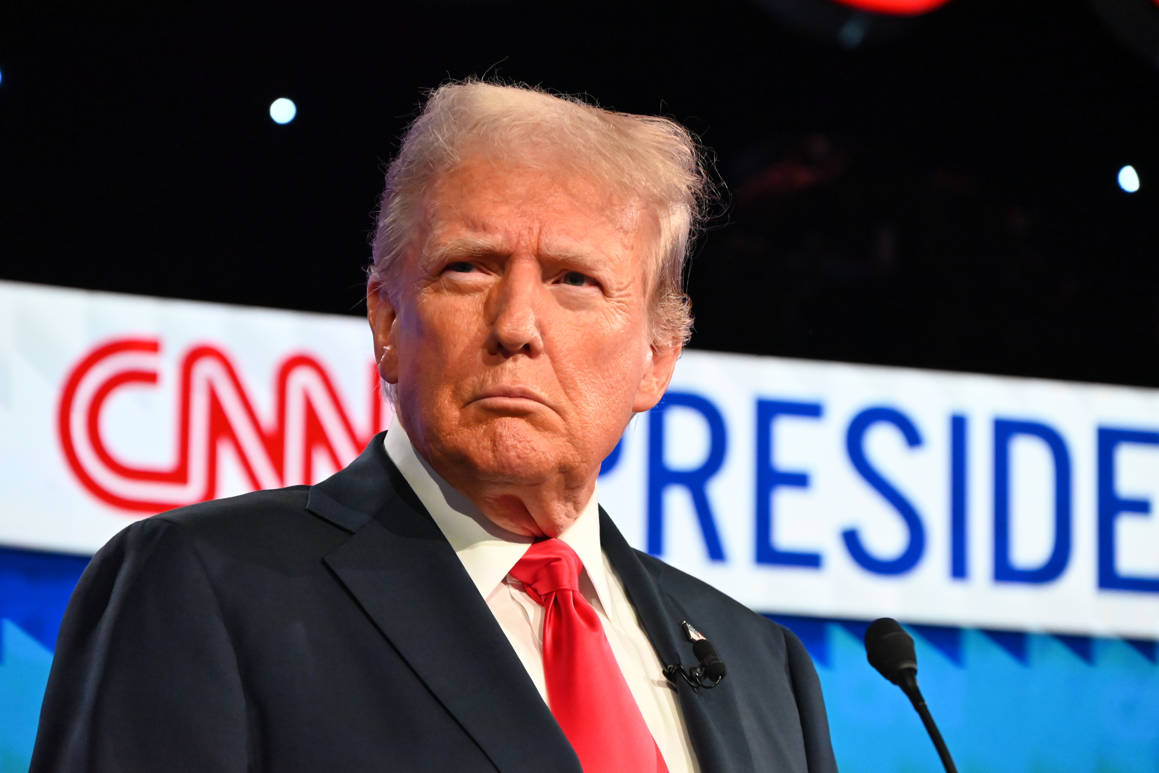Donald Trump stands in front of a "CNN President" sign, wearing a suit with a red tie, at a televised event