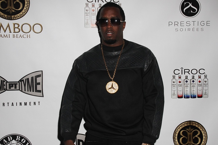 Sean "Diddy" Combs poses on the red carpet in a stylish, dark outfit with a large pendant necklace and sunglasses, in front of a backdrop featuring various logos