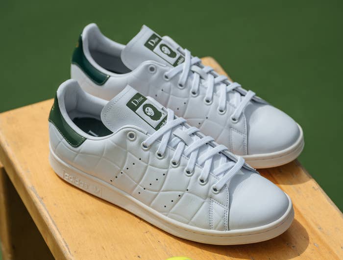 White Adidas Stan Smith sneakers with green accents are displayed on a wooden bench, next to a yellow tennis ball on a court