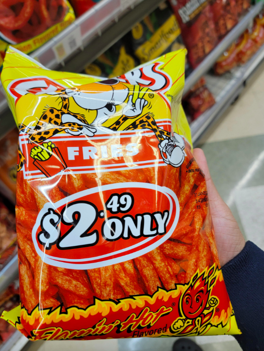 A hand holding a bag of Flamin' Hot Fries snack, priced at $2.49 only, in a grocery aisle with other snack bags in the background