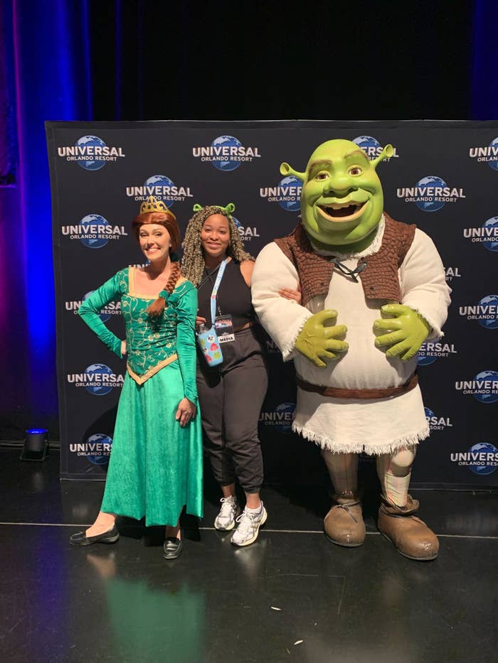 Princess Fiona, a woman posing between them, and Shrek are at a Universal Orlando Resort event backdrop. Fiona is in medieval attire and Shrek is in his typical outfit