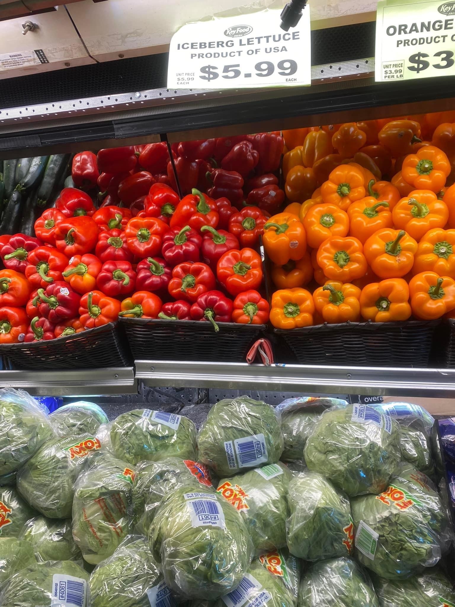 Grocery store produce section displaying red and orange bell peppers on upper shelves and packaged iceberg lettuce below, with visible price signs