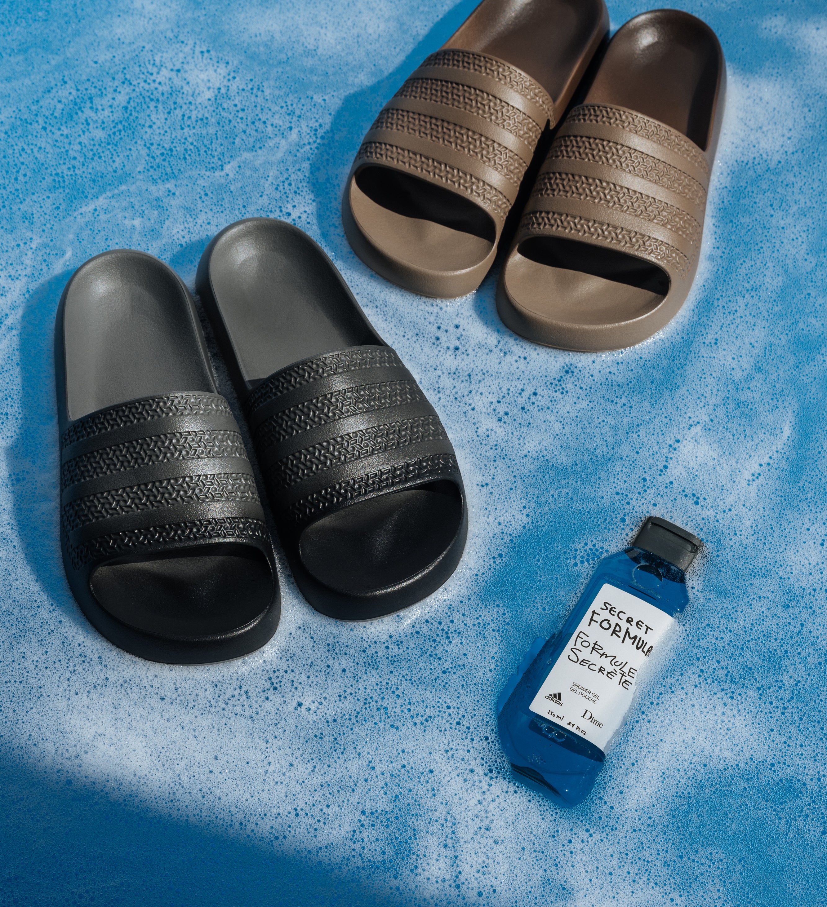 Four pairs of Adidas slides in shades of gray and brown are positioned on a textured blue surface, next to a bottle labeled &quot;SCETP for People Sickle.&quot;