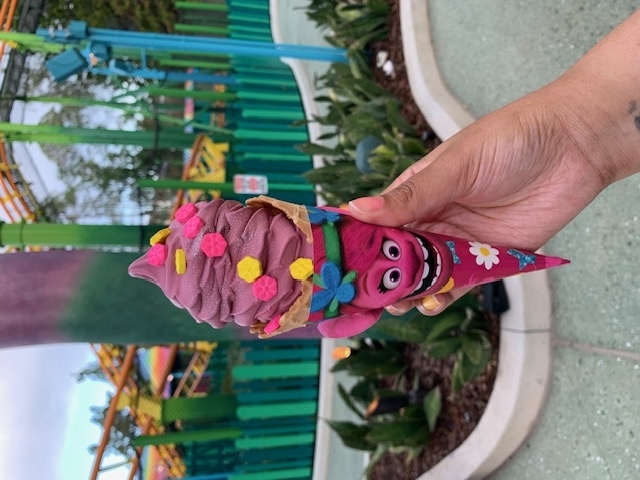 A hand holds a pink-themed ice cream cone shaped like Poppy from "Trolls," against a theme park backdrop featuring roller coaster tracks and greenery