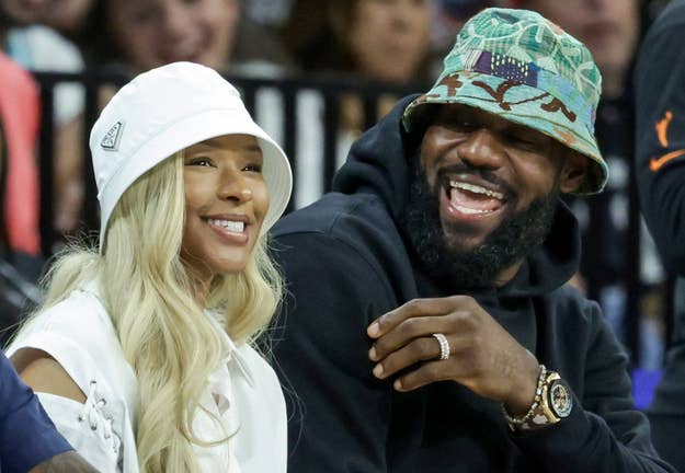 Savannah James and LeBron James, both wearing bucket hats, smiling and sitting together at an event. LeBron wears a watch and a patterned hat; Savannah's is plain