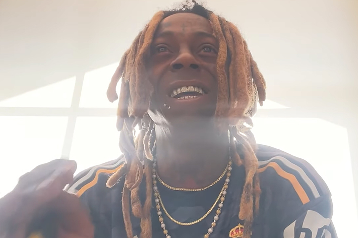 Lil Wayne smiling, wearing a sports jersey with gold necklaces and his signature dreadlocks