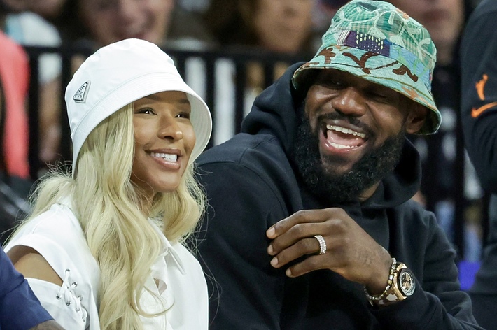 LeBron James and Savannah James are smiling while seated at an event. Both are wearing bucket hats; Savannah wears a white outfit, and LeBron has a dark jacket