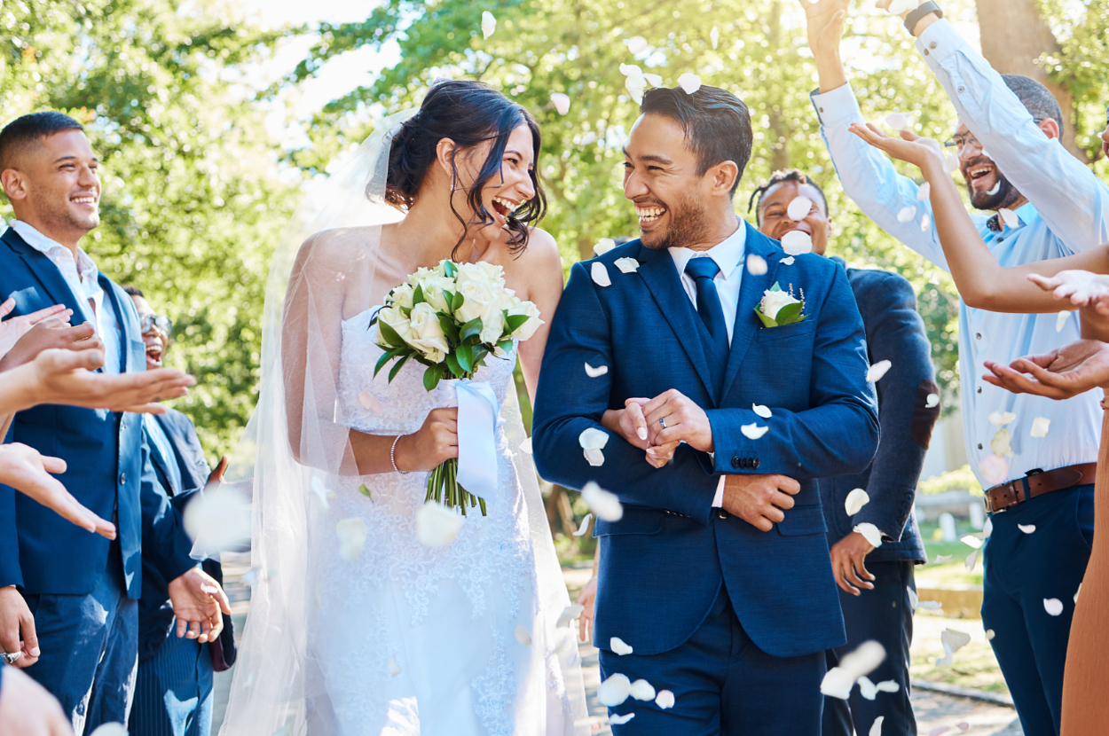 Bride and groom in wedding attire walk through crowd celebrating with flower petals. The bride holds a bouquet and the couple smiles joyfully