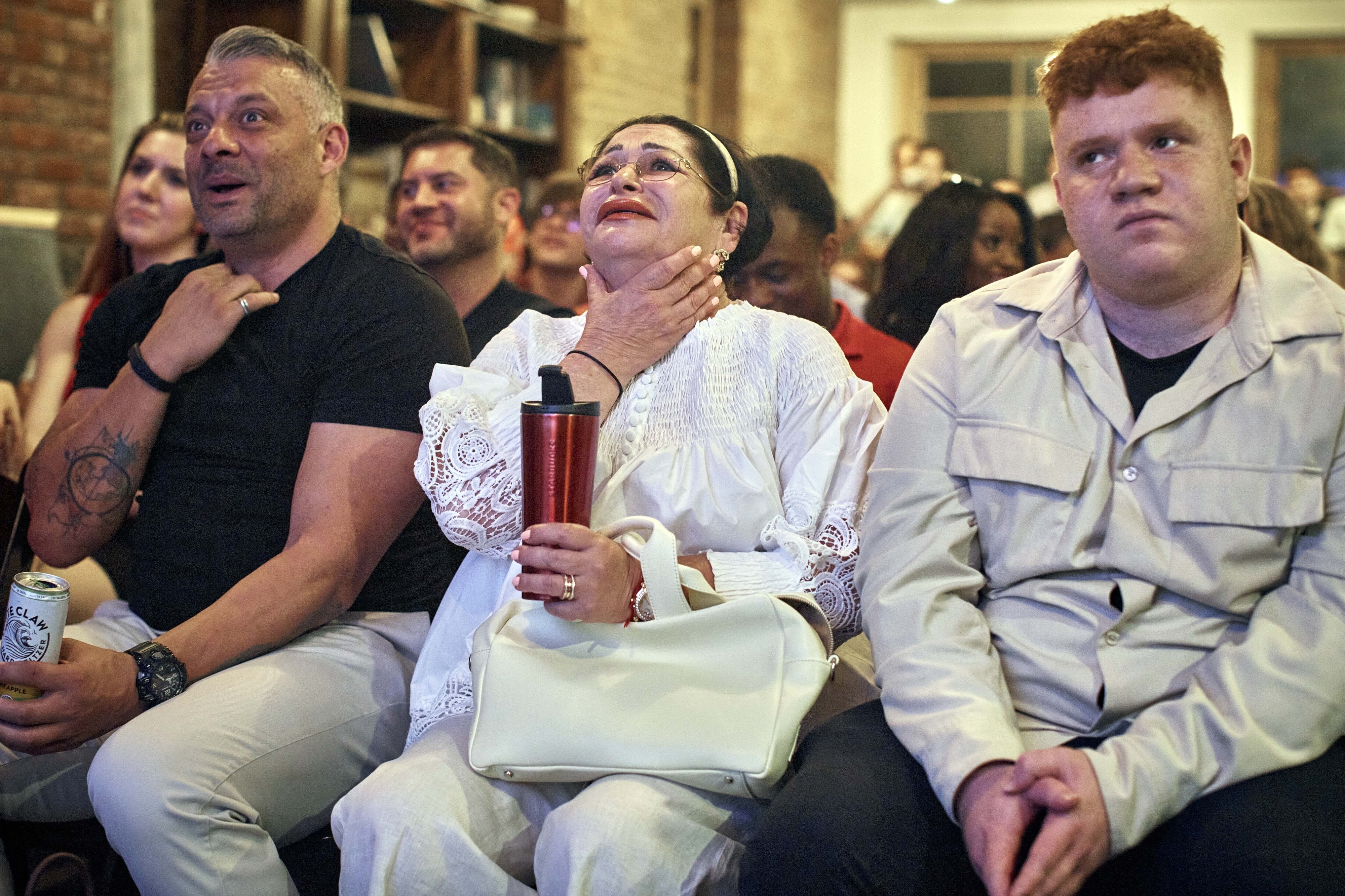 Three people are seated in a crowd with varied expressions: one looking surprised, one looking emotional, and one looking indifferent. Names not known