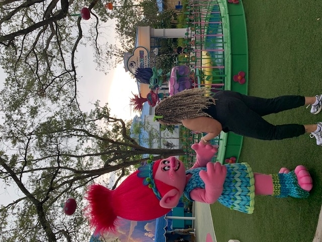 A person interacts with Poppy, a pink character from the Trolls movie, in an amusement park setting. Poppy wears a blue dress