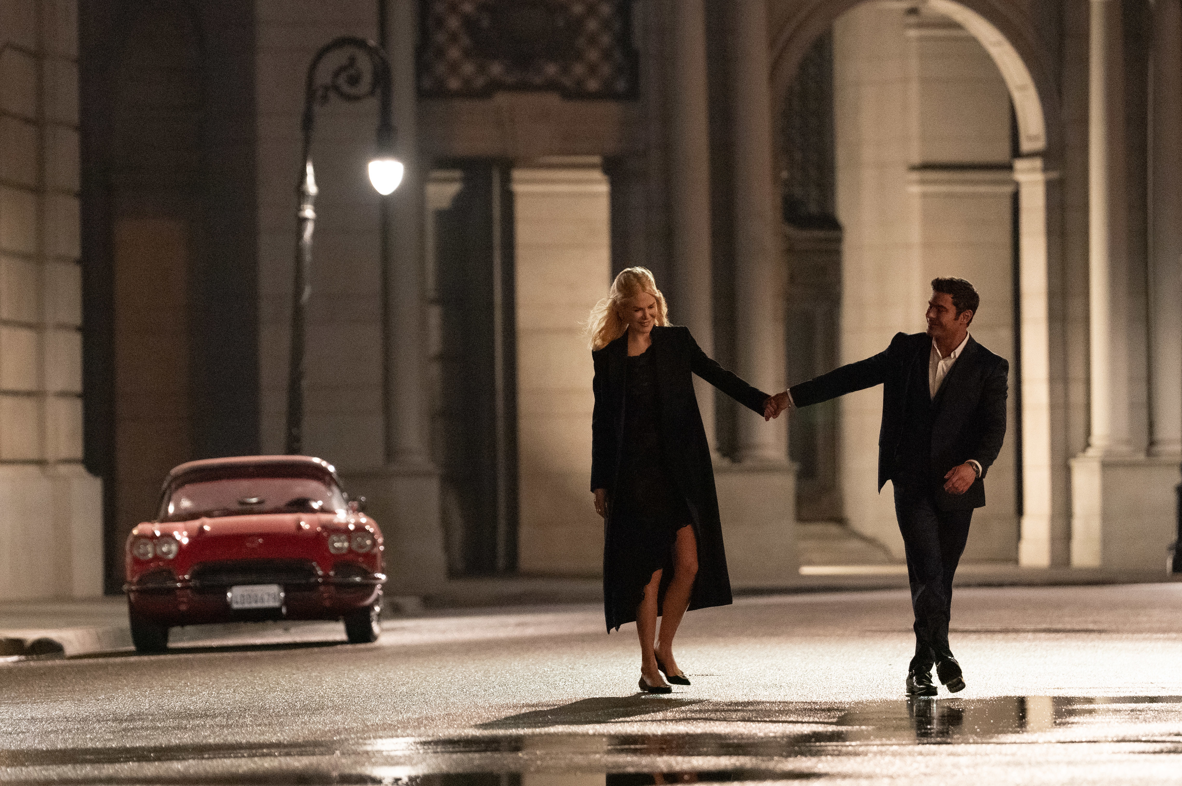 Context from the article suggests the image features Nicole Kidman and Zac Efron. Both are walking and holding hands at night in an elegant urban setting