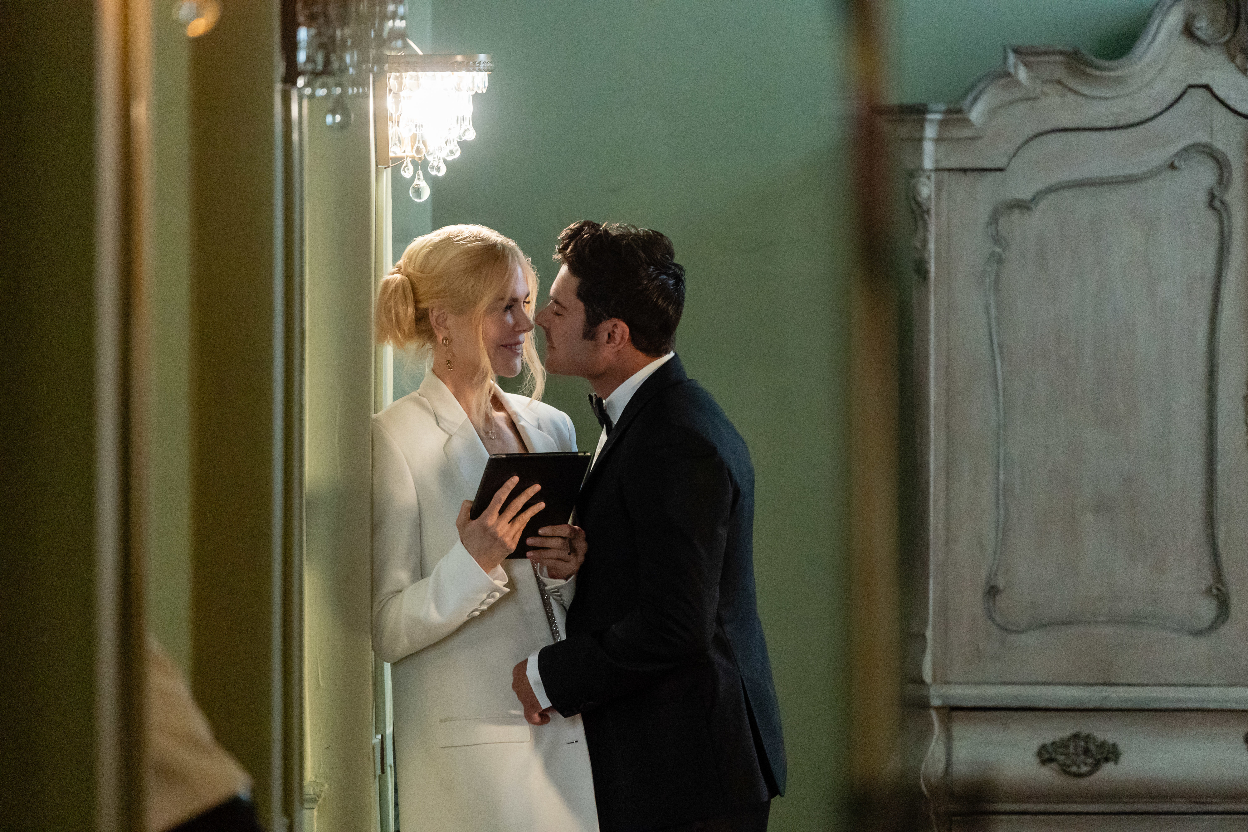 Nicole Kidman and Zac Efron are elegantly dressed, sharing an intimate moment in a refined room. Foster holds a small book, and Latif leans in to kiss her