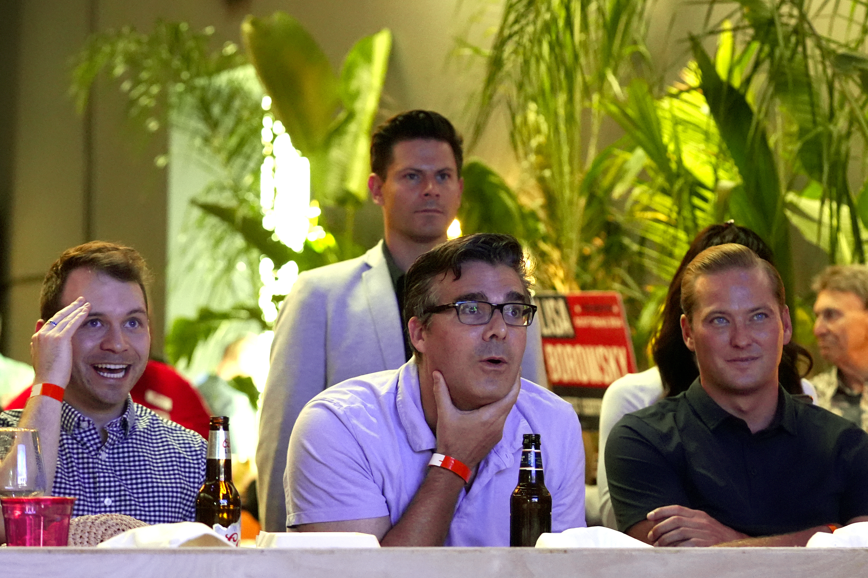 Four men at an event reacting to something off-camera with varying expressions. Unidentified bottles and drinks are on the table in front of them