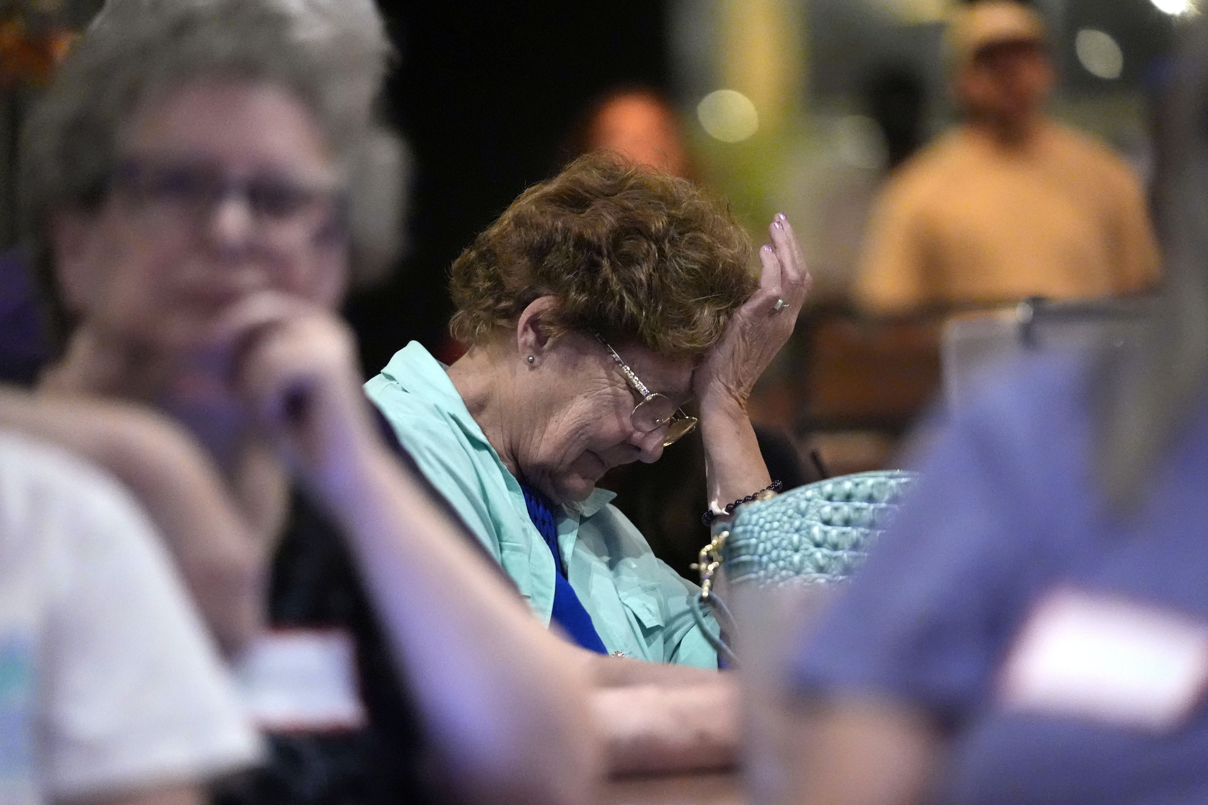 Several people seated in a dimly lit room, with one elderly woman in the foreground holding her head in her hand, appearing distressed